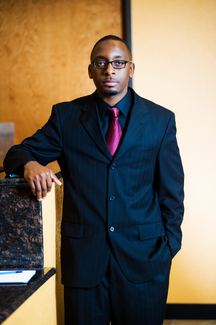 Professional portrait of a clinic administrator wearing a dark pinstripe suit, black shirt, and maroon tie standing next to the reception counter